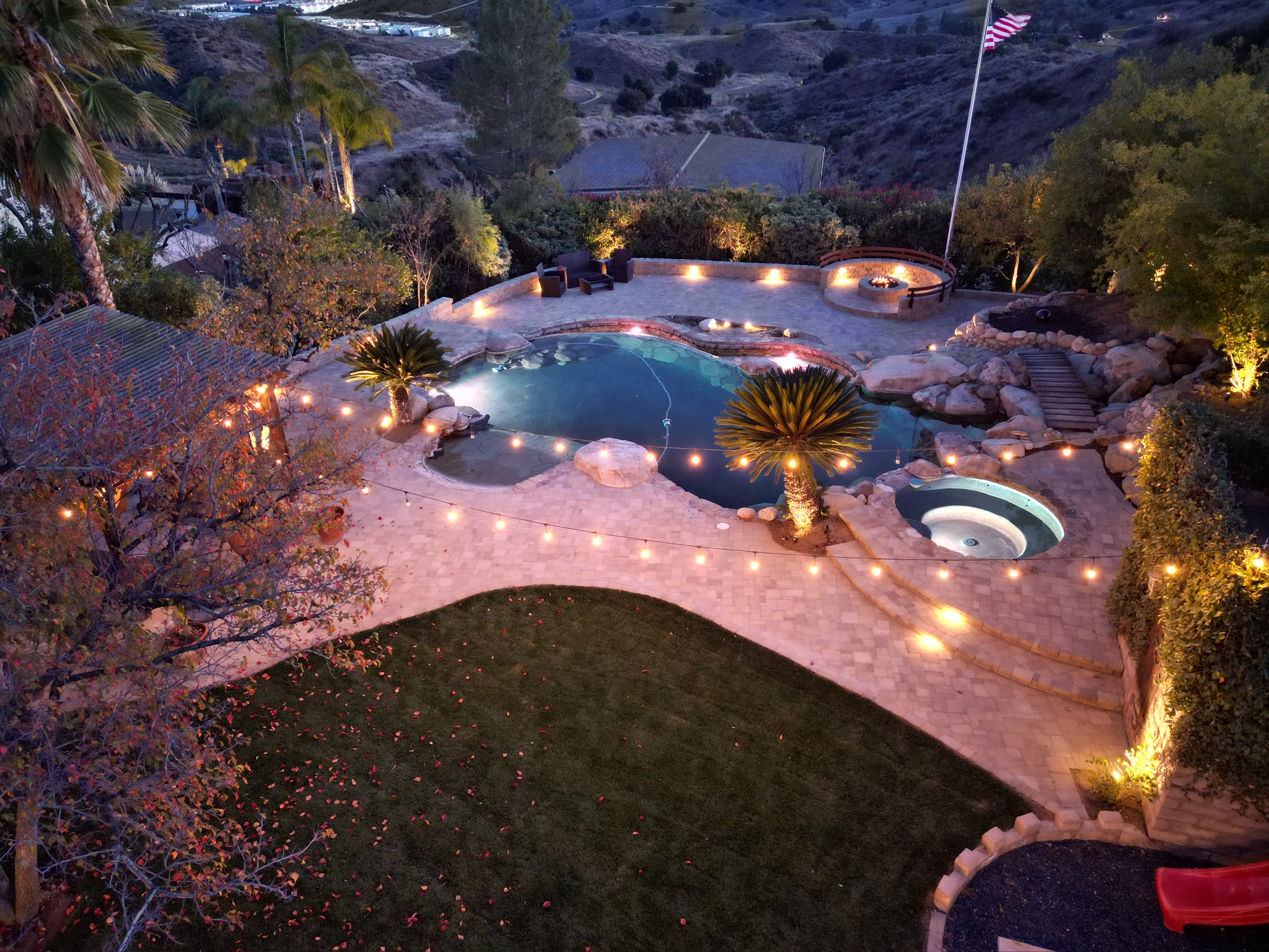 Full backyard with pool, fire pit, and string lights — drone shot at dusk